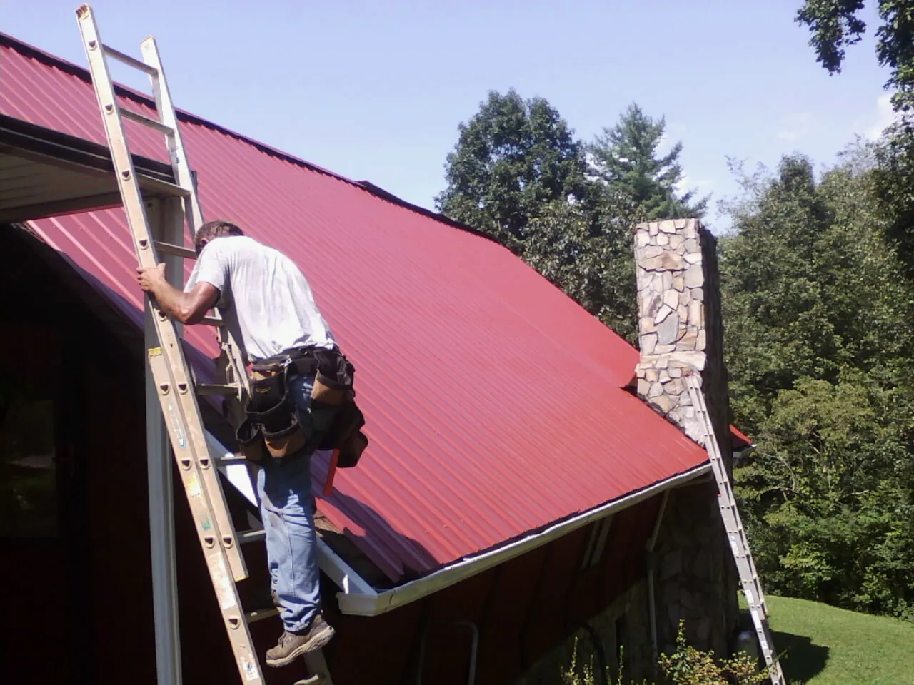 Roofer on a ladder beside a red metal roof and stone chimney — Soffit & Fascia Repair by Roofing Brothers in Chadwick